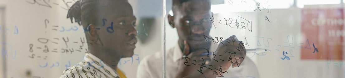 Two male students wearing white shirts writing on a transparent glass board solving a math equation in a well-lit classroom.