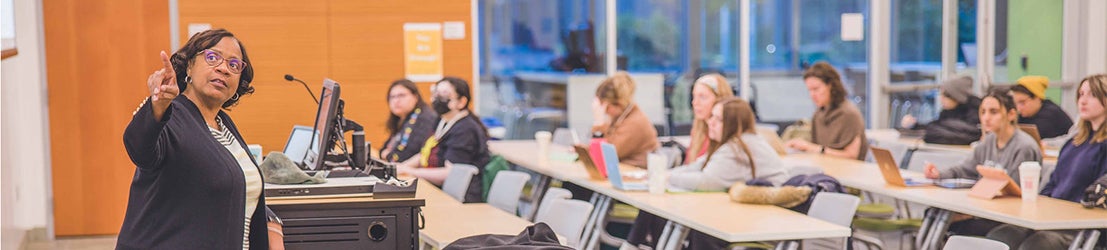 A female teacher pointing at a whiteboard in front of students in a classroom.