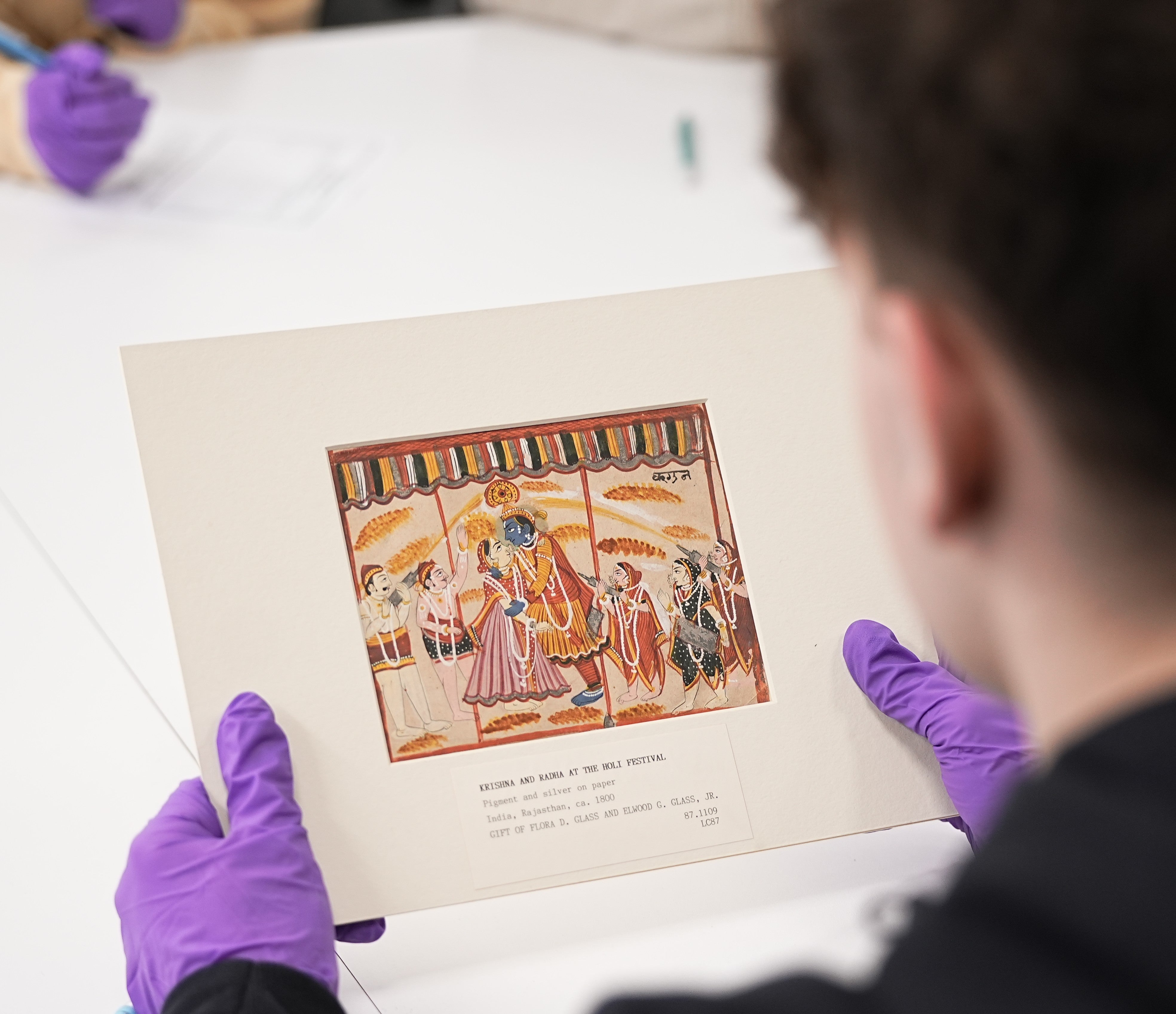 A person wearing purple gloves examines an image of Krishna and Radha from the CMA Education Art Collection at a table during a group activity.