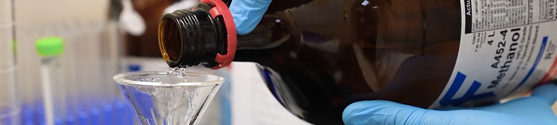 A student with blue gloves pour chemical liquids into a flask.