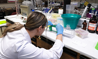 A student in a white lab coat working on research in a lab.