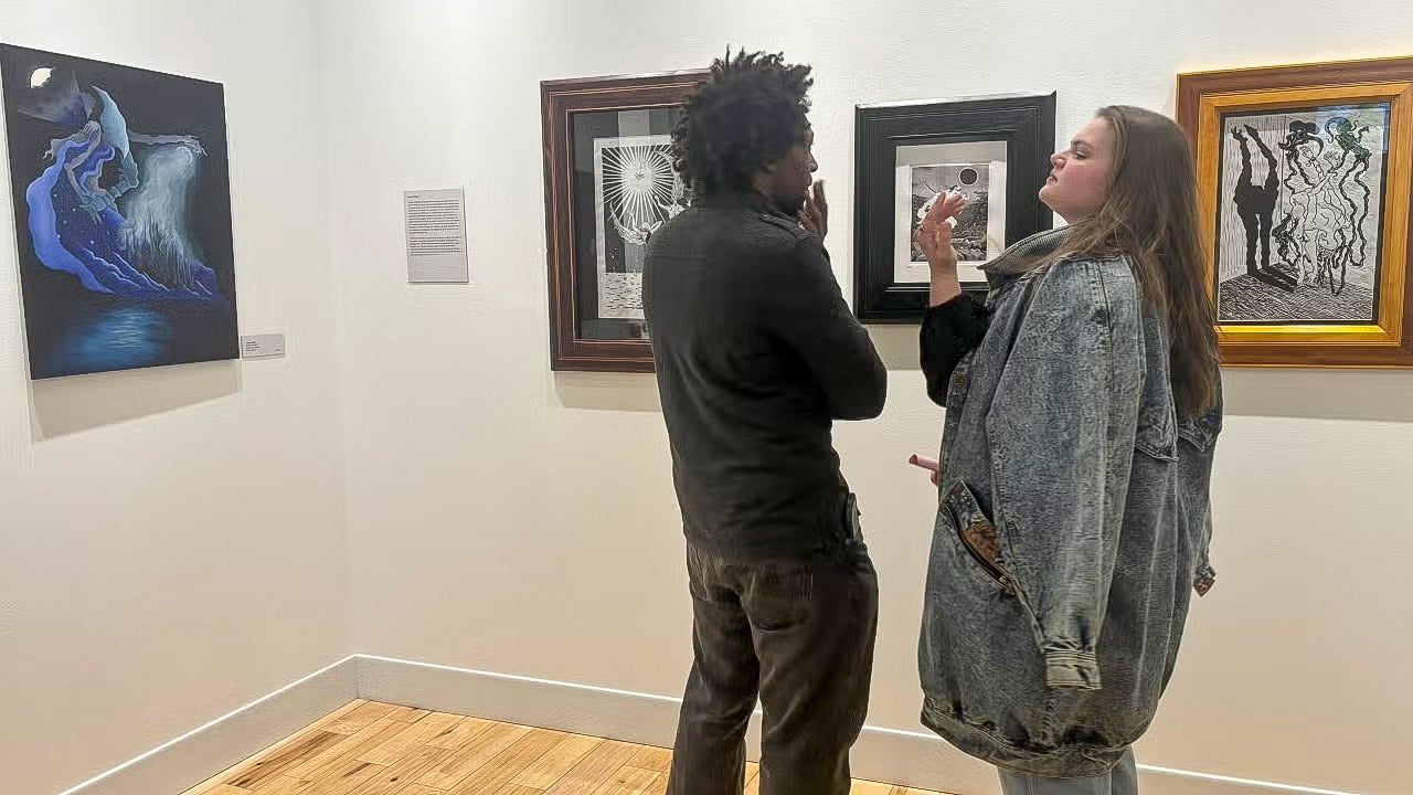 Two students talk in front of student artworks installed in an art gallery.