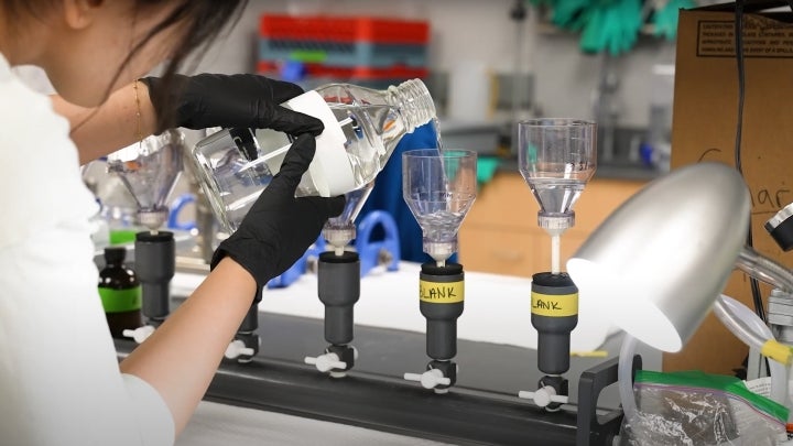 An EVS student in a lab coat with black gloves pouring chemical solution into a container in a lab.