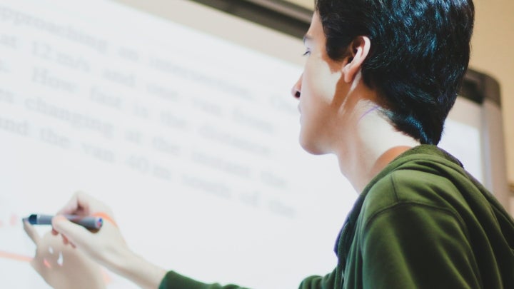 A female student in a green sweater writing and solving a math equation on a smart whiteboard in a classroom.