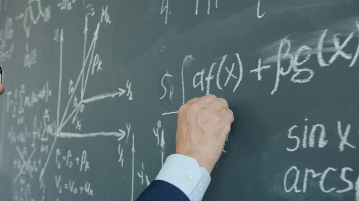A male professor writing a math equation with chalk on a green chalkboard.