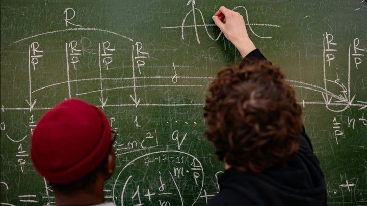 A male student closely observing a professor graphing a math equation on green chalk board.