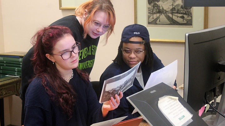 A group of three people review historical documents together at a desk with a computer and printed materials.