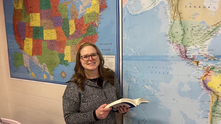Ohio History Teacher of the Year Marilyn Orseno stands in front of large world and U.S. maps while holding an open book. 