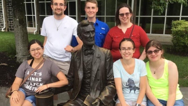 A group of people sitting and standing around a bronze statue of a seated figure on an outdoor bench, with trees and a building in the background.