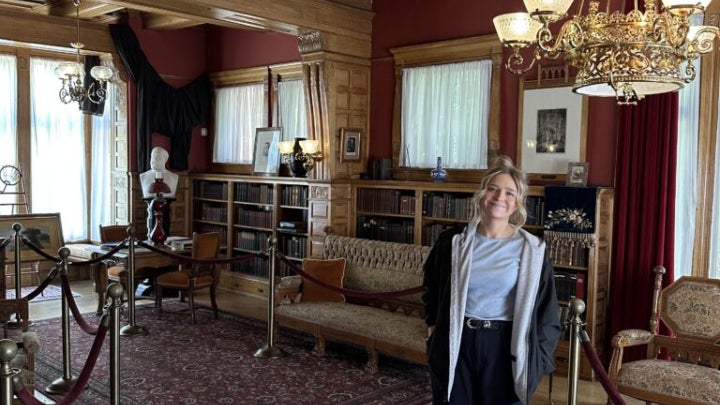 Person standing inside an ornate historic room with wood-paneled ceilings, chandeliers, bookshelves, and antique furniture at the Garfield NPS site.
