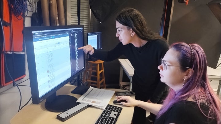 Two interns work at a computer workstation, with one pointing at a large monitor while the other uses a keyboard in a studio setting.