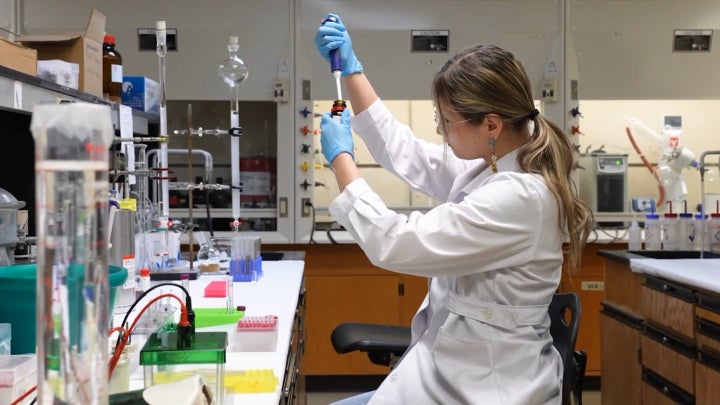 A student in a white lab coat and blue gloves working on research in a lab.
