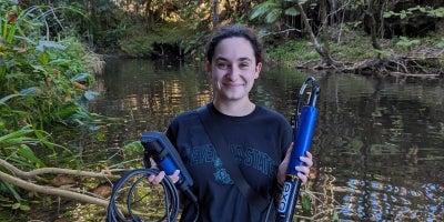 A student holding EVS equipment for field research outside next to a river.