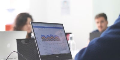 A student in a blue sweater working on graphs on a laptop along with two other students in a classroom.