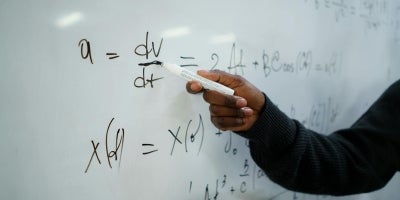 A male student wearing a black sweater pointing at a math equation with a dry erase marker on a whiteboard in a classroom.