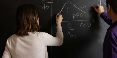 A female student and a male professor graphing a math equation on a black chalkboard.