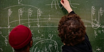 A male student closely observing a professor graphing a math equation on green chalk board.