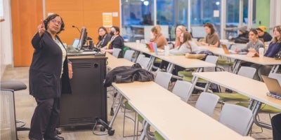 A female teacher pointing at a whiteboard in front of students in a classroom.