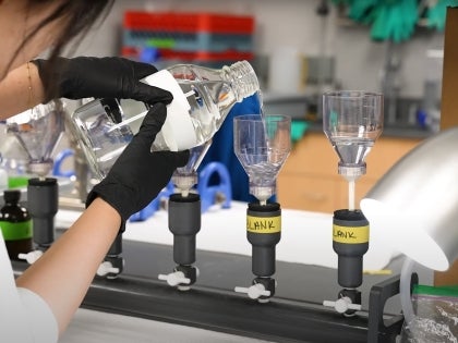 An EVS student in a lab coat with black gloves pouring chemical solution into a container in a lab.
