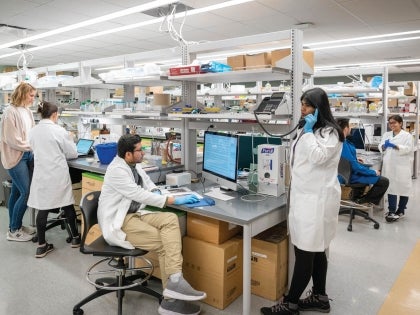 Two BIO students in lab coats in busy lab facility conducting research.