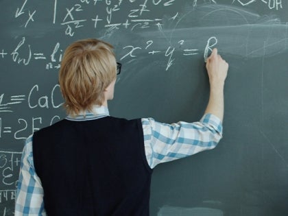 A male professor writing on a green chalkboard writing a math equation with chalk.