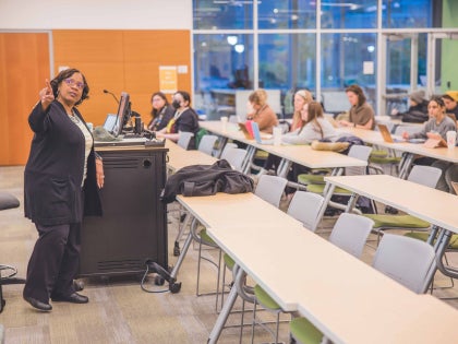 A female teacher pointing at a whiteboard in front of students in a classroom.