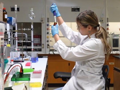 A student in a white lab coat and blue gloves working on research in a lab.