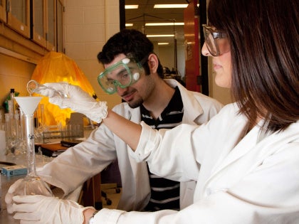 Two students in lab coats and lab goggles handling research equipment in a lab. 