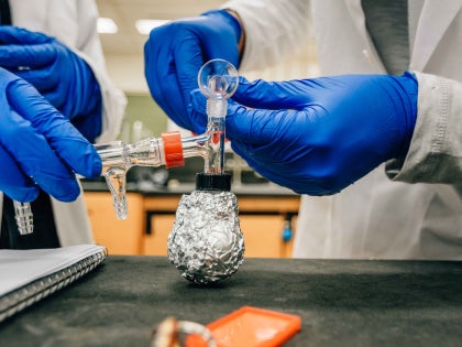 Two students a in white lab coats and blue gloves pouring chemical liquids into a flask in a lab.